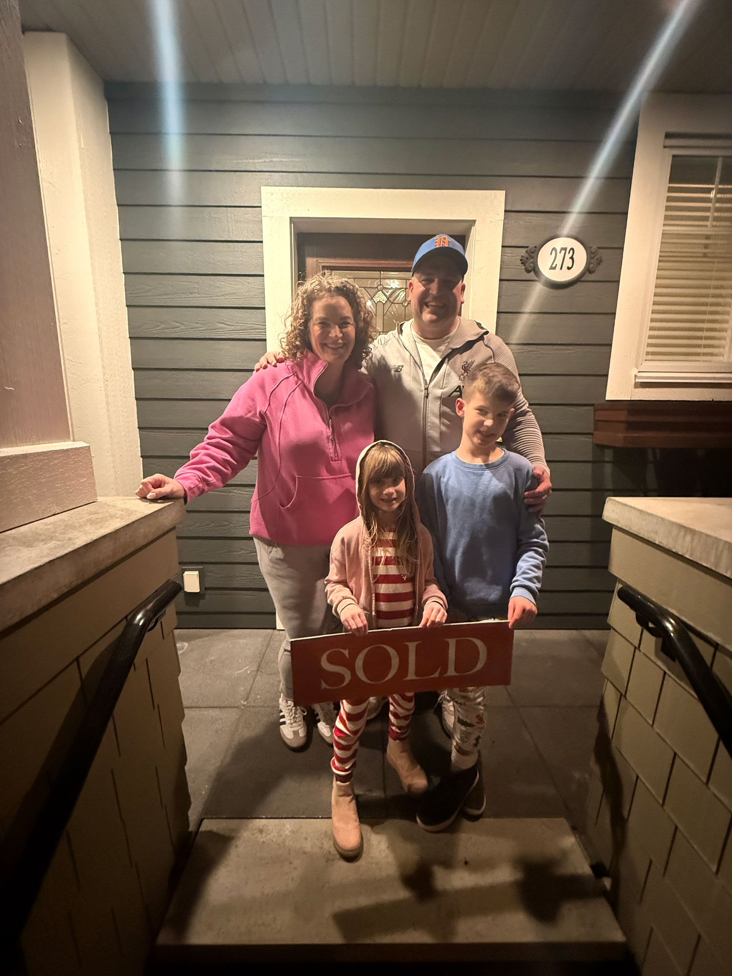 A happy family of four stands on the front steps of their new home at night, holding a 'SOLD' sign and celebrating their successful purchase.