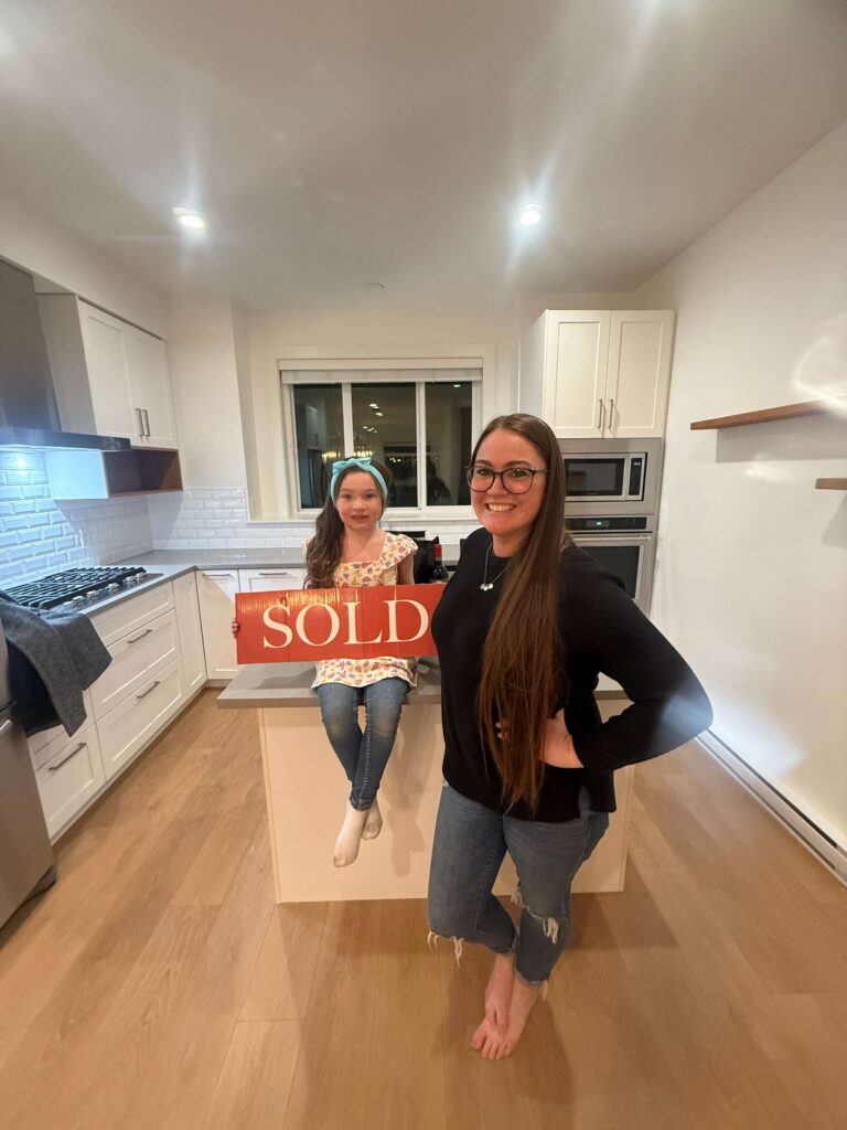 A smiling mother and daughter celebrating their new home in a bright, modern kitchen. The daughter, sitting on the kitchen island, holds a bold red 'SOLD' sign—a perfect symbol of patience, persistence, and the dream home they manifested.