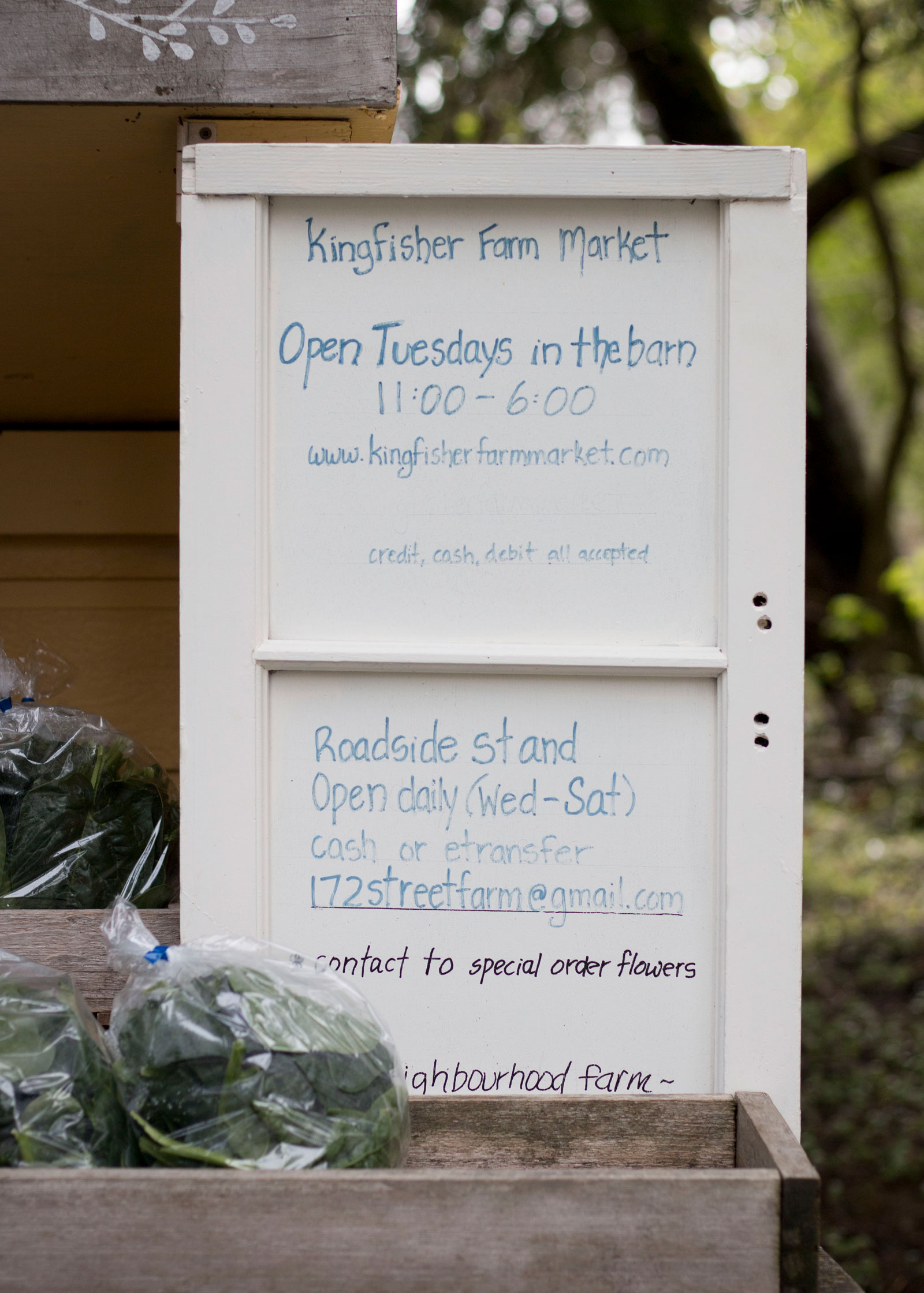 A hand-painted sign at Kingfisher Farm Market displaying opening hours and payment options, with fresh, locally grown produce in wooden crates nearby.